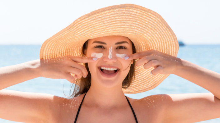 An image of a woman smiling, she has a wide-brimmed hat on her head and sunscreen across her face, she is at the beach.